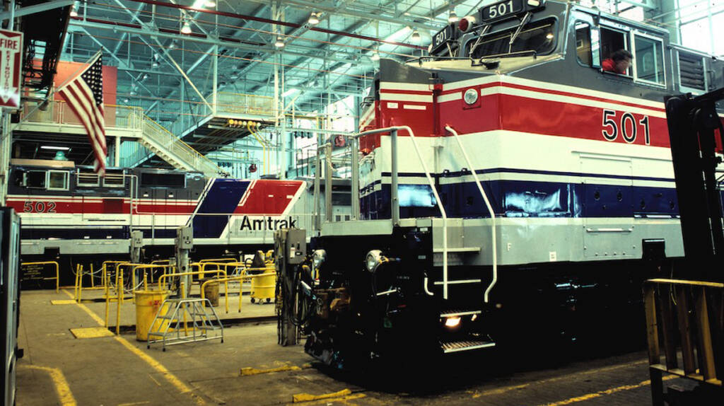 A gray locomotive with wide red, white, and blue stripes sits in a large and brightly lit factory building. A similar engine is in the background.