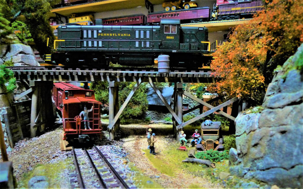 A red toy train passes under a bridge carrying another toy train on a toy train layout