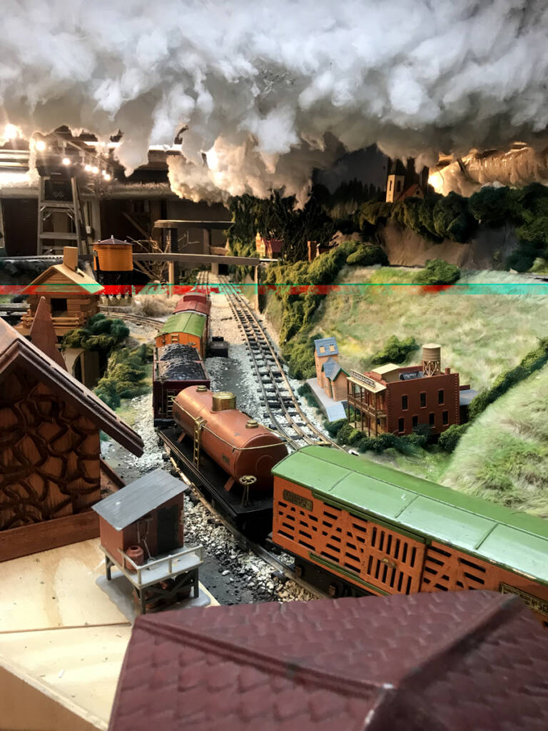 A standard gauge toy train on a toy train layout with modeled clouds in the sky above the layout