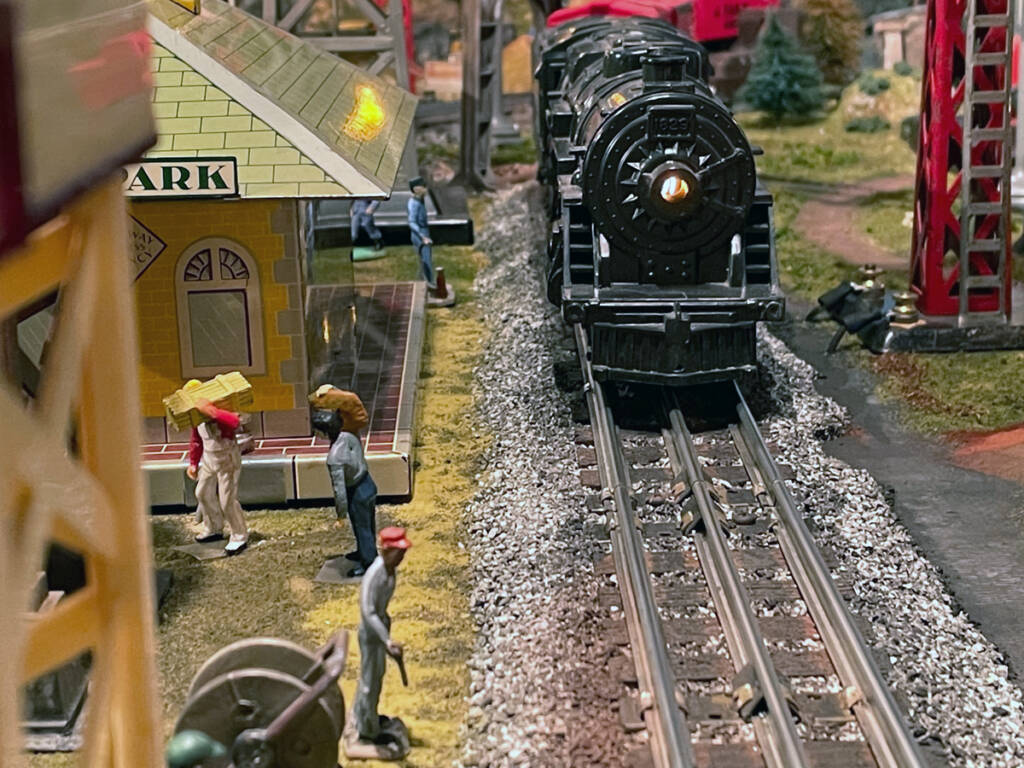 A steam locomotive leads a toy train into a passenger station on a toy train layout