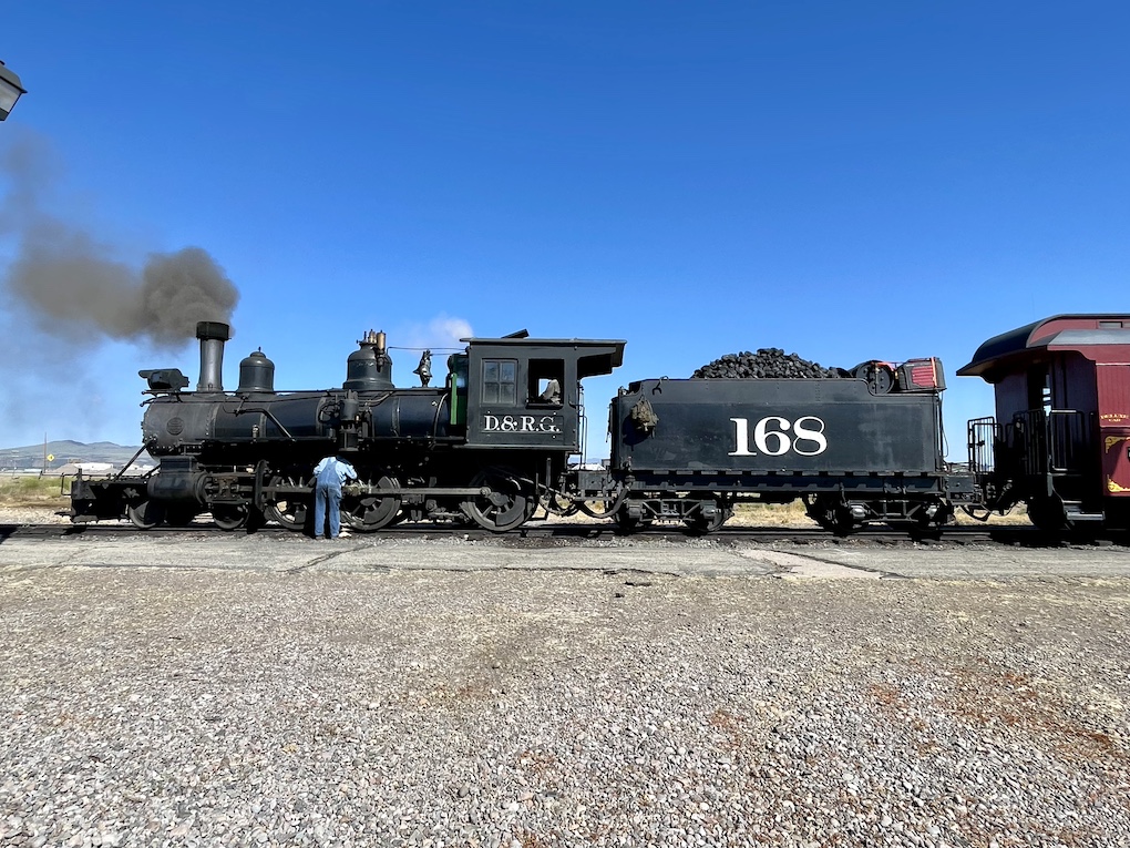 Sideview of an old steam locomotive being looked over by a crewman wearing bib overalls