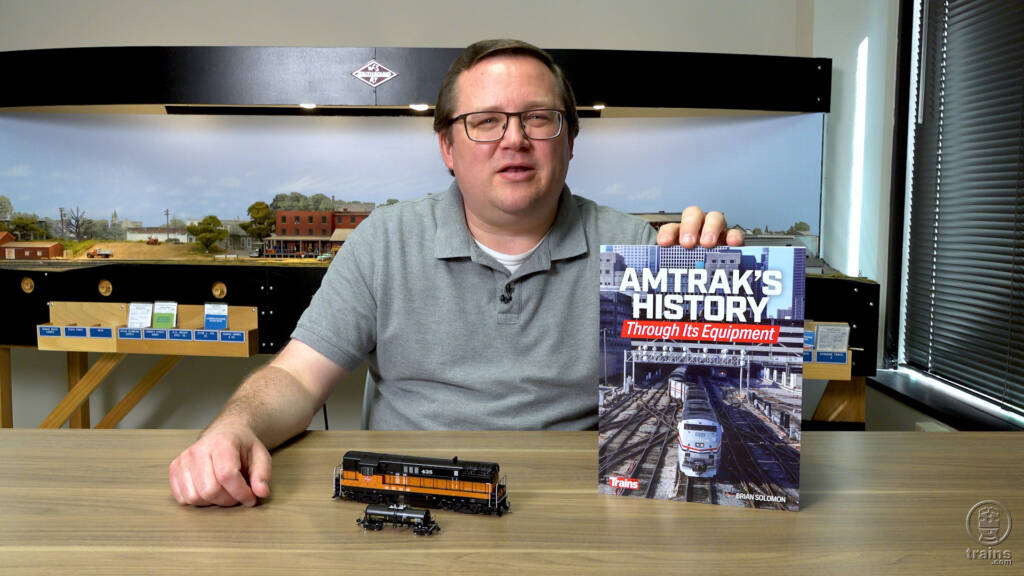 Image of man with brown hair, glasses, and gray shirt holding a book with two model trains in foreground and HO scale layout in background.