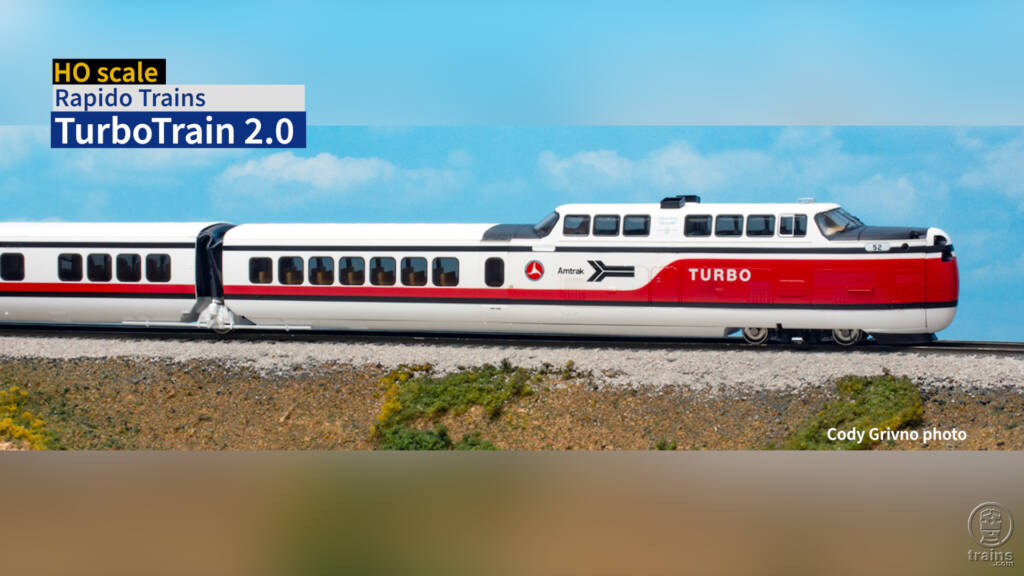 Color image of man with goatee wearing glasses and blue shirt standing in front of an HO scale model railroad with trains and structures. Title bar is visible in upper left corner.