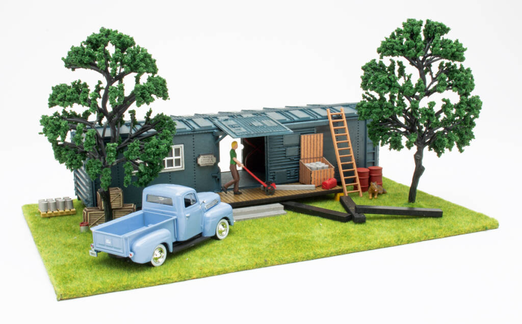 Color photo of O scale rail maintenance office with boxcar body, pickup truck, trees, and figures on scenicked base on white background. 