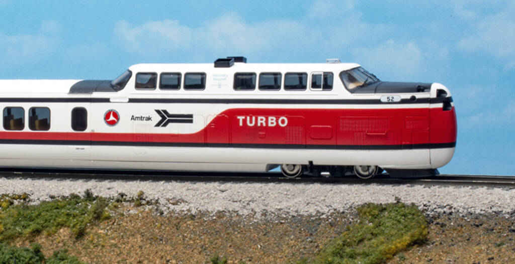 Futuristic toy train painted in red, white, and black colors on a model base with green grass and a tan concrete culvert in front of a blue sky backdrop with white fluffy clouds.
