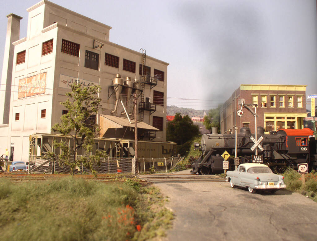 A car sit in the foreground in front of a model train and a model factory structure