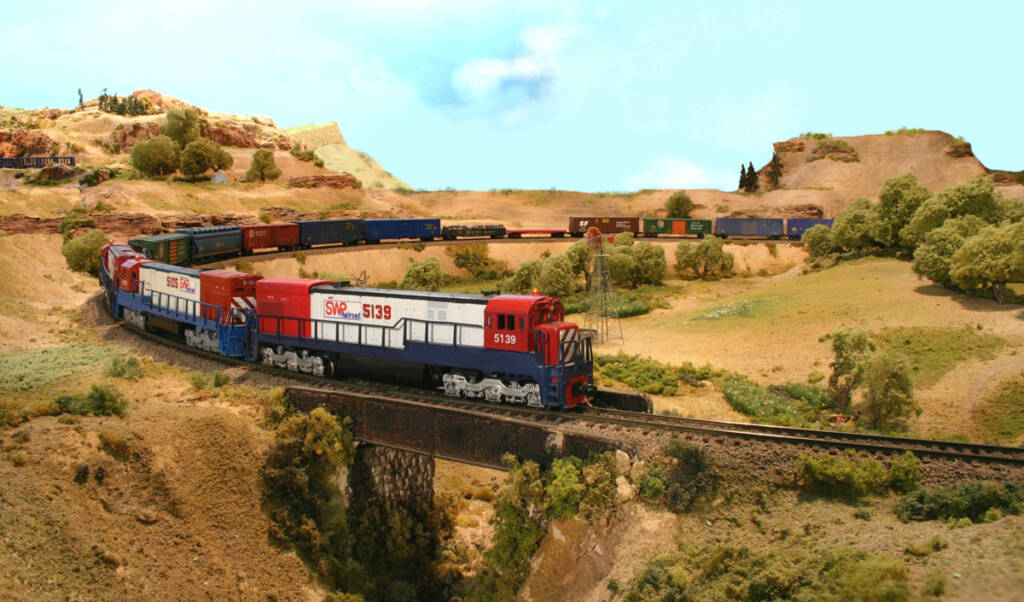 A model freight train is lead by a black, white, and red model locomotive over a bridge in a desert setting
