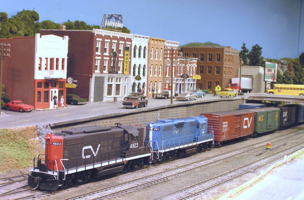 A model freight train is lead by a black and red model locomotive on a model railroad layout with vehicles and a town in the background
