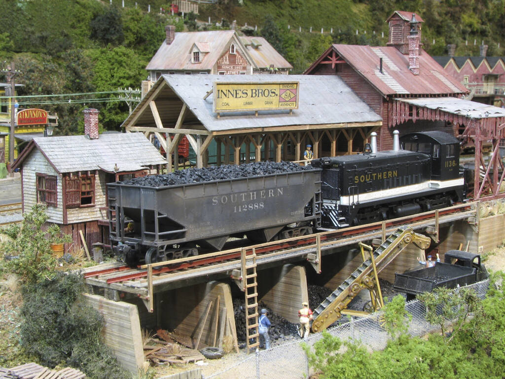 A black diesel locomotive and a black freight car sit at a coal and lumber industrial stop on a model railroad layout