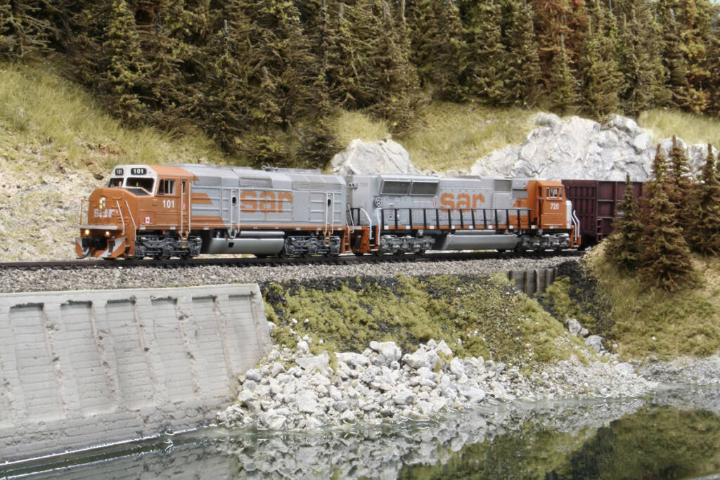 Two gray and orange model locomotives lead a freight train past a body of water on a model railroad layout