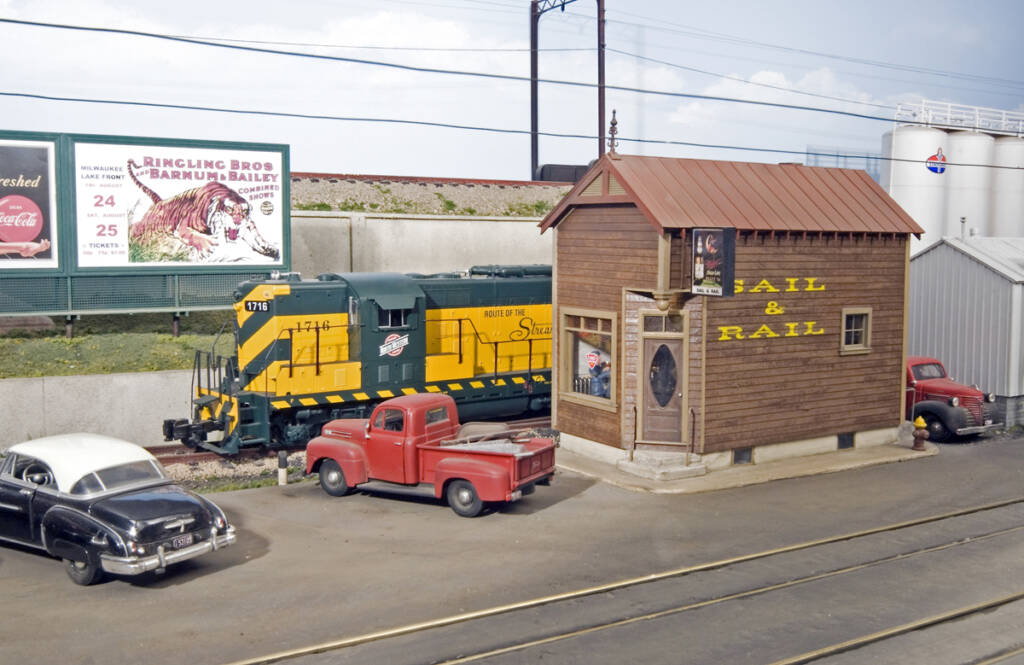A model train passes a bar with letting on the side of the bar which reads "The Sail & Rail" on a model railroad layout