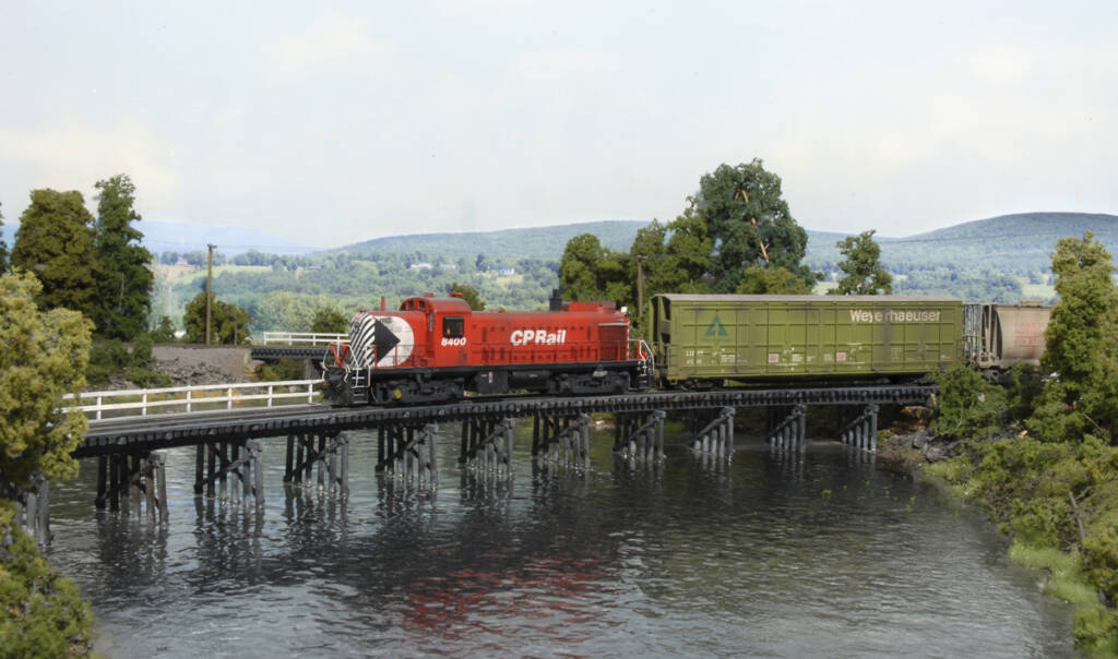 A red model caboose brings up the rear on a model train as it crosses a river on a model railroad layout
