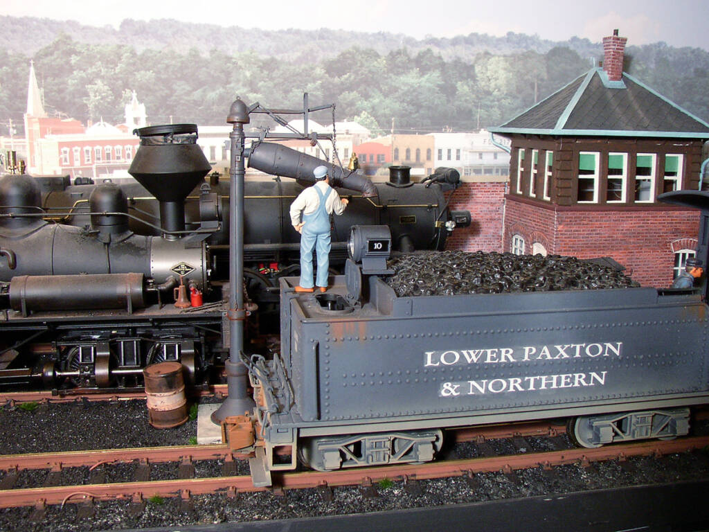 A model figure stands on a steam locomotive tender on a model railroad layout