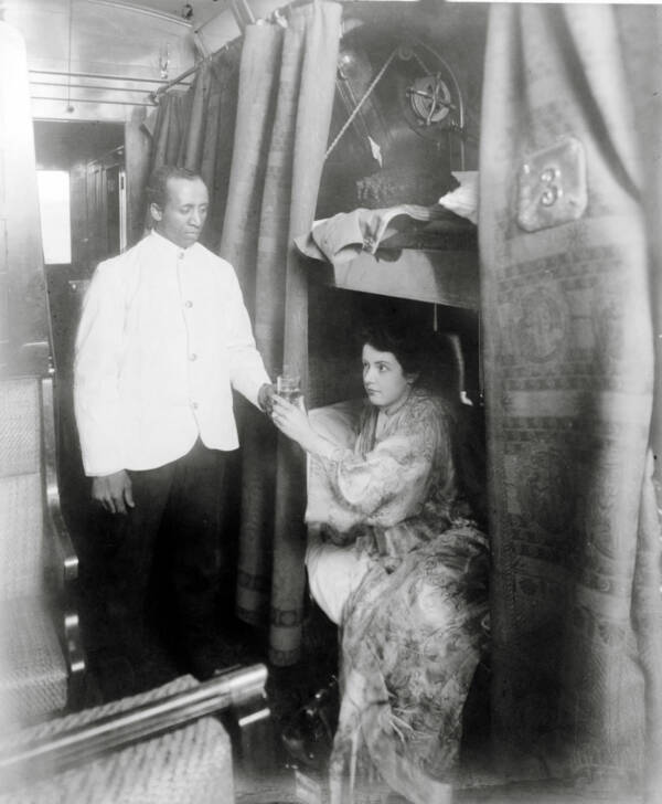 A Pullman Porter serves a glass of water to a woman in a sleeping car berth