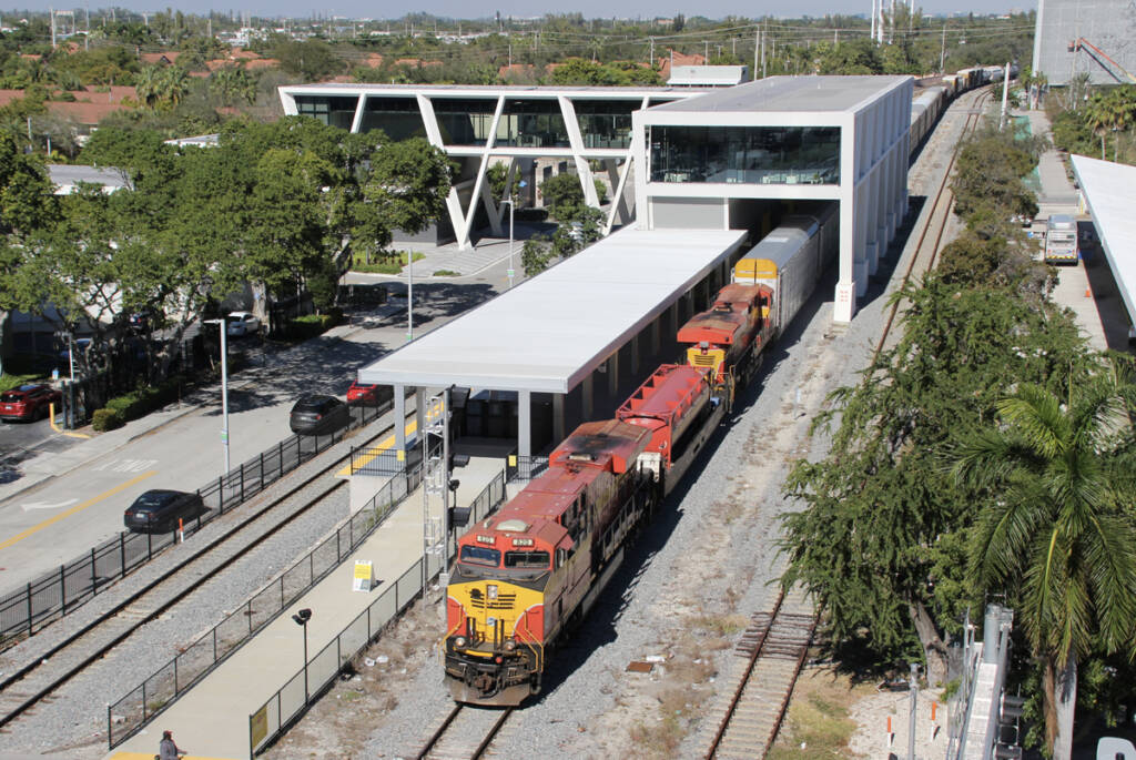 Freight train with orange and yellow locomotives passes through passenger station