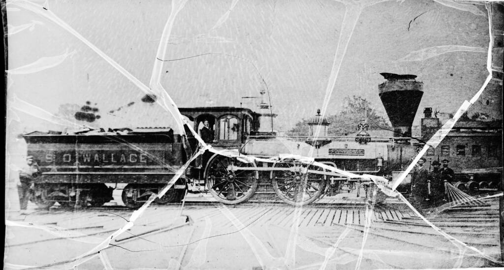 A side view of an early steam locomotive taken on a glass plate negative. A man is in the cab and three railroad workers stand at the front of the engine
