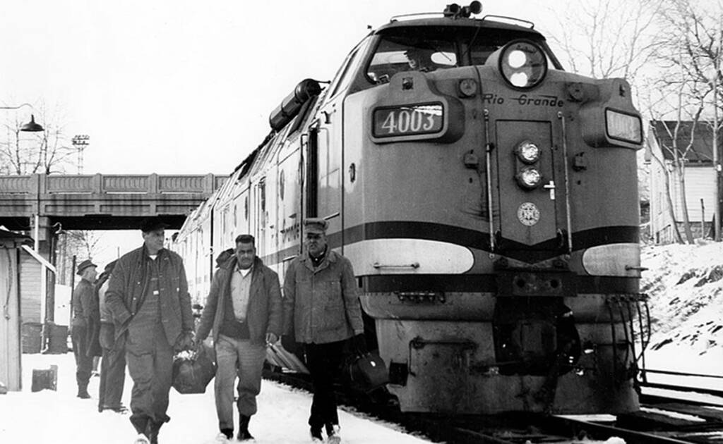 A black and white phot of three men walking past a locomotive