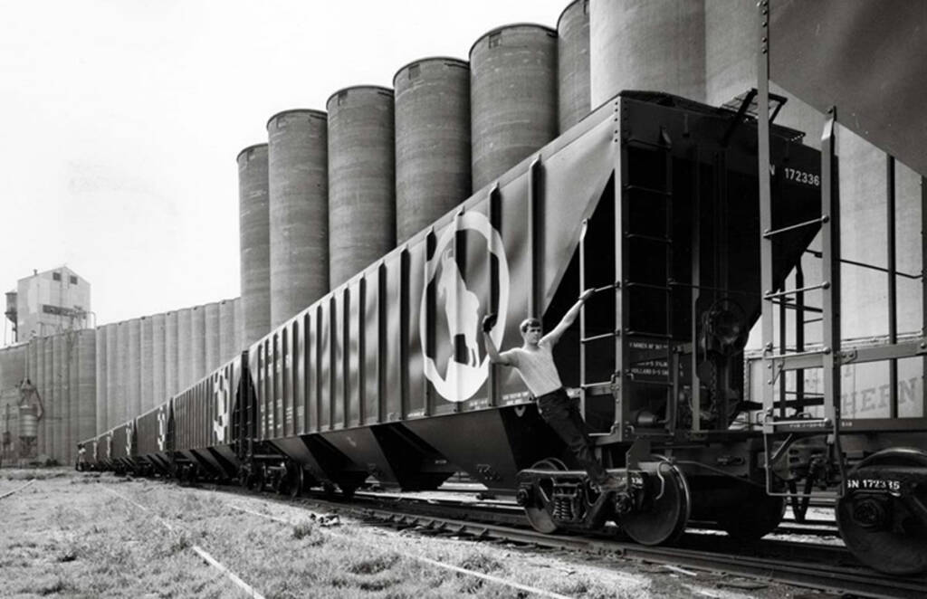 A man hangs off the side of a grain hopper as it advances through an industrial yard in a black and white photo