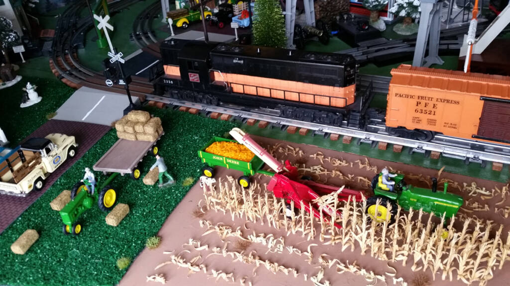 A black and orange toy locomotive leads a train past a farm with a tractor on a toy train layout