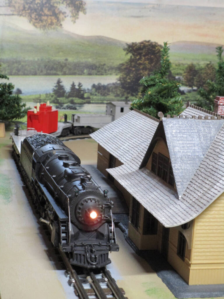 A black steam locomotive passes a passenger station on a toy train layout
