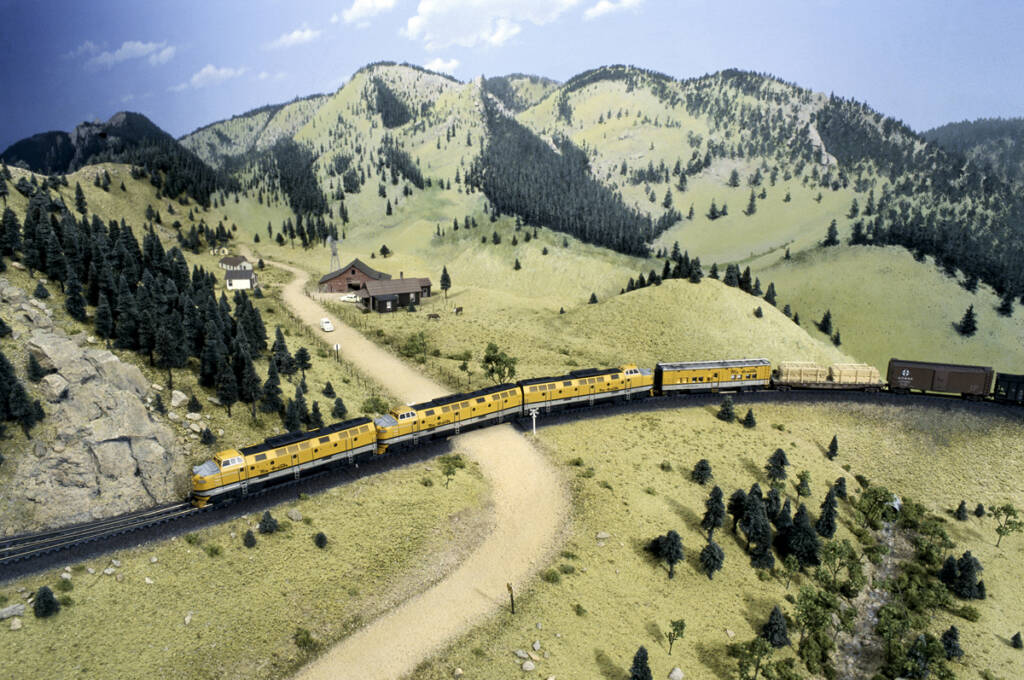 A consist of three yellow model locomotives leads a freight train through a mountainous setting on a model railroad layout