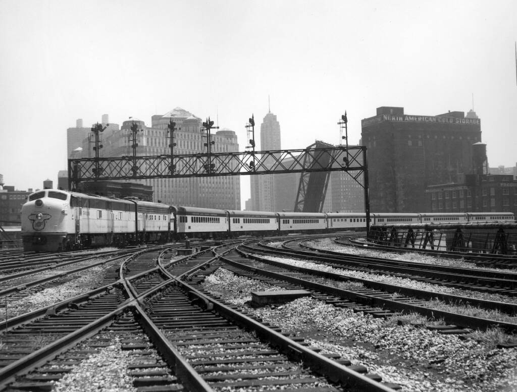 Black-and-white photo of streamlined passenger train departing a large city