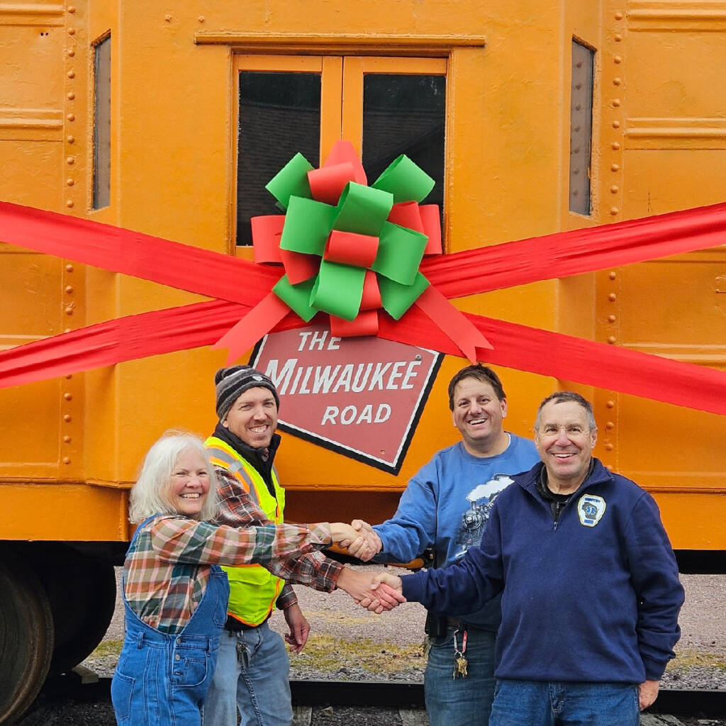 Four people shaking hands next to an orange caboose decorated with a Christmas ribbon. Milwaukee Road caboose donated.