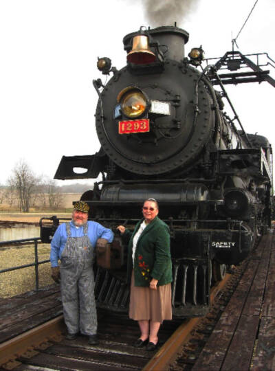 Man and woman standing on turntable in front of steam locomotive. Laura Jacobson, museum co-founder, dies.