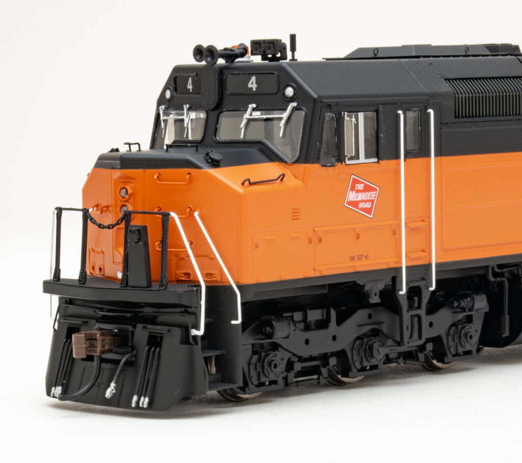Color photo showing nose and fireman’s side of HO scale six-axle cowl-body diesel painted orange and black on white background.