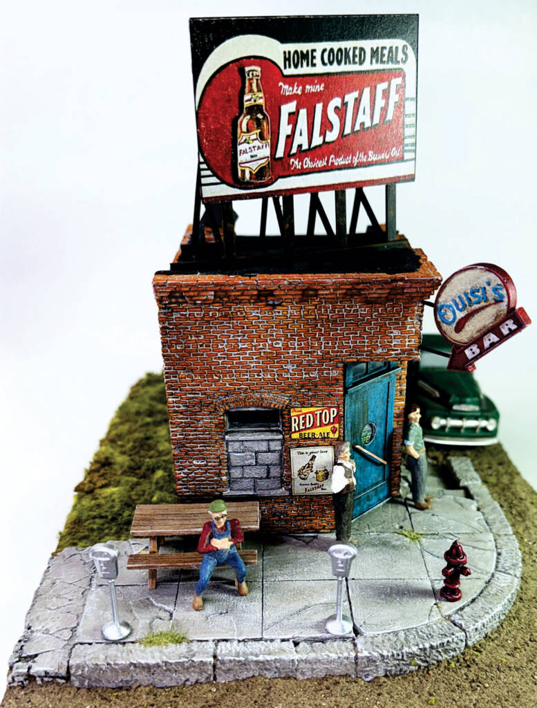 Color photo of O scale brick corner building with teal door, billboard on roof, sign on corner, figures, vehicle, and other details on scenicked base with white background.