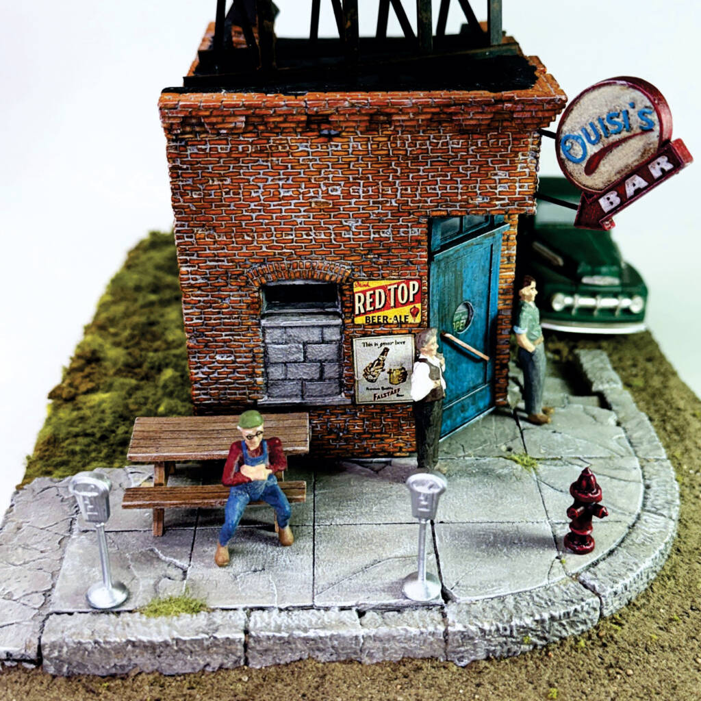 Color photo of O scale brick corner building with teal door, billboard on roof, sign on corner, figures, vehicle, and other details on scenicked base with white background.