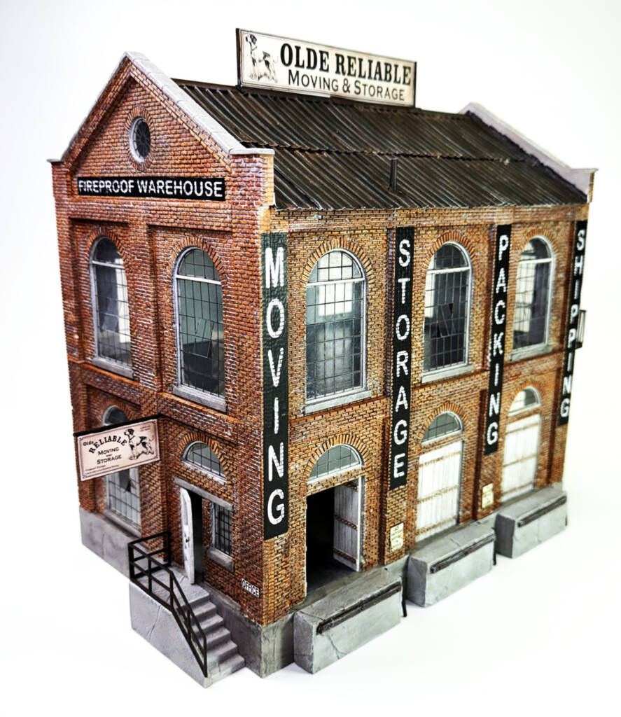 Color photo of O scale two-story brick warehouse with peaked roof, black-and-white signs, large windows, and roll-up doors on white background.