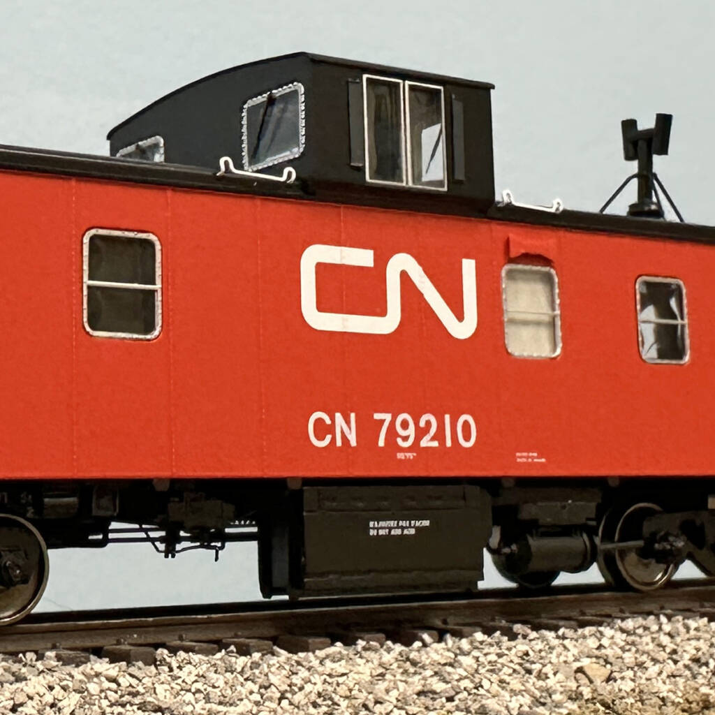 Color photo showing part of HO scale caboose painted red and black with white graphics on scenicked base with a blue backdrop.