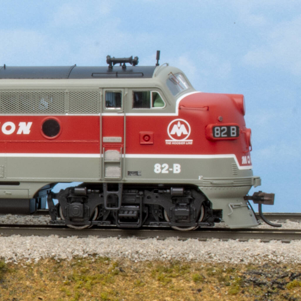 Color photo showing engineer's side of HO scale cab unit painted red, gray, black, and white on scenicked base with a sky blue backdrop.