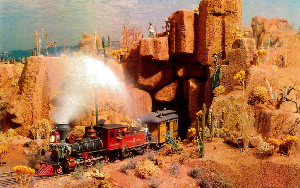 Color photo of red and black steam locomotive pulling yellow passenger car with black roof through Desert Southwest scenery.
