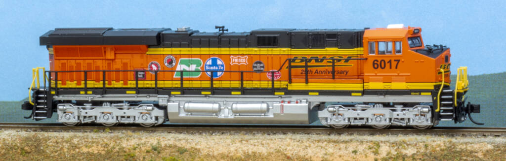 Color photo of modern N scale diesel locomotive painted orange, black, and silver with various heralds on the long hood on a scenicked base with a sky blue background. 