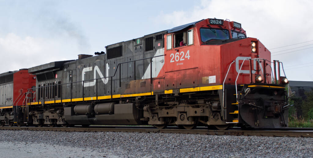 Color photo of modern GE six-axle road diesel painted red, white, and black with yellow sill stripe. A barn is partially visible in the background.
