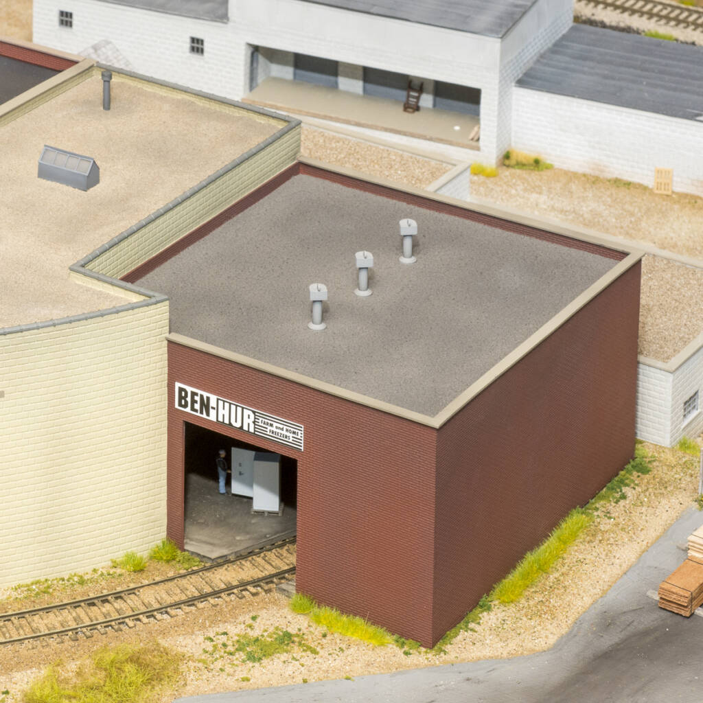 Color photo of tan and brown brick warehouse buildings with gravel roof on scenicked model railroad layout with a cinder block building in the background.