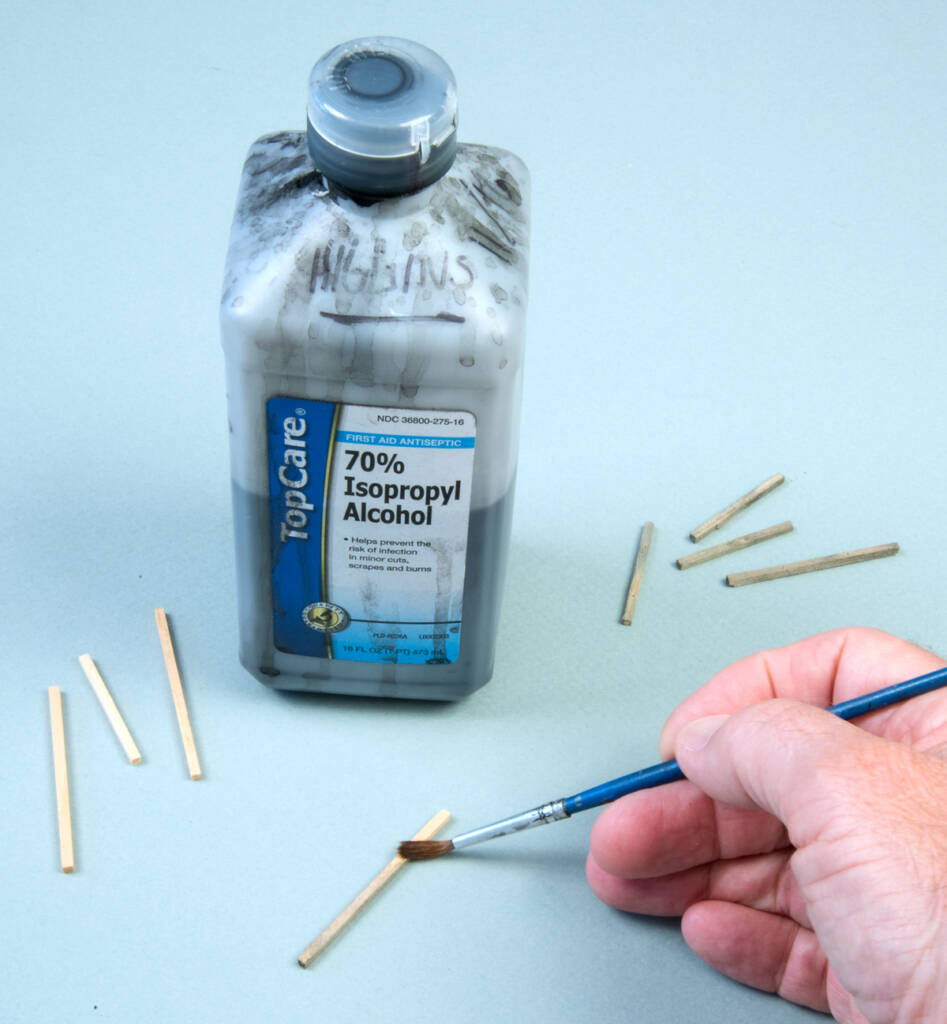 Color photo of a man’s right hand holding a paintbrush with blue handle applying stain to pieces of stripwood on a light blue background. A bottle of isopropyl alcohol with a blue, white, and gold label with black printing is also visible. 