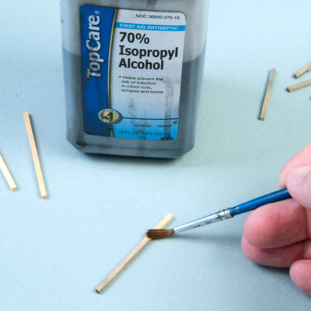 Color photo of a man’s right hand holding a paintbrush with blue handle applying stain to pieces of stripwood on a light blue background. A bottle of isopropyl alcohol with a blue, white, and gold label with black printing is also visible.