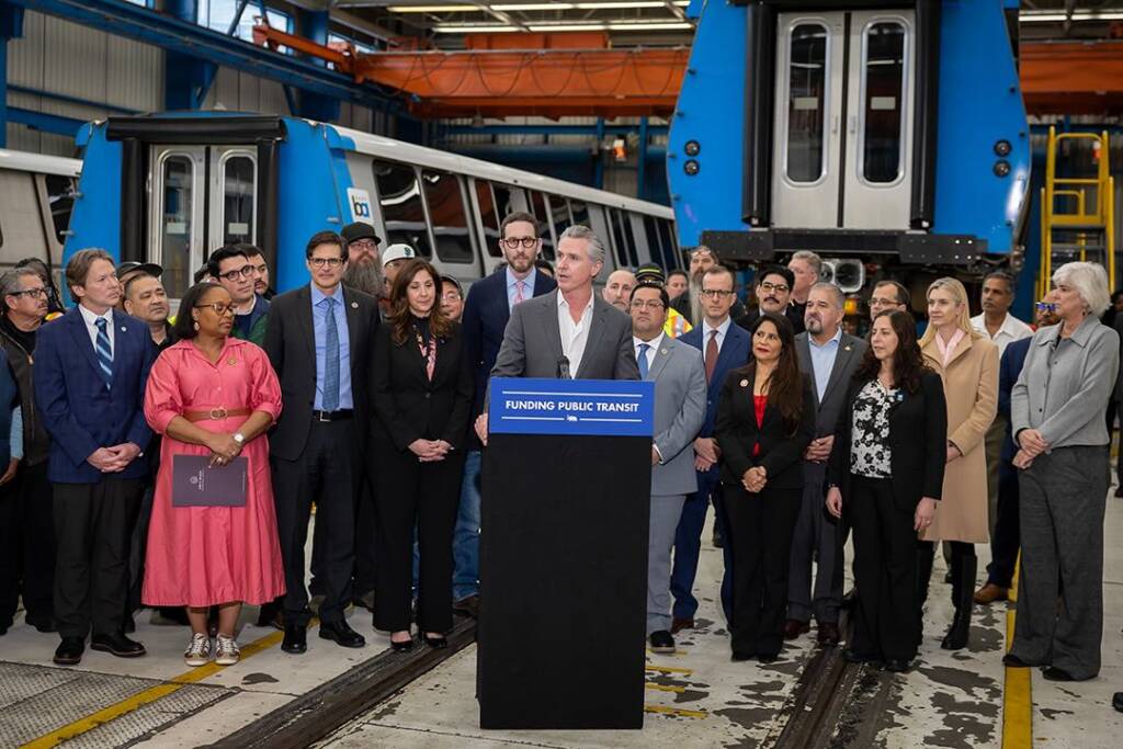 Man speaking at podium while surrounded by people in transit maintenance facility