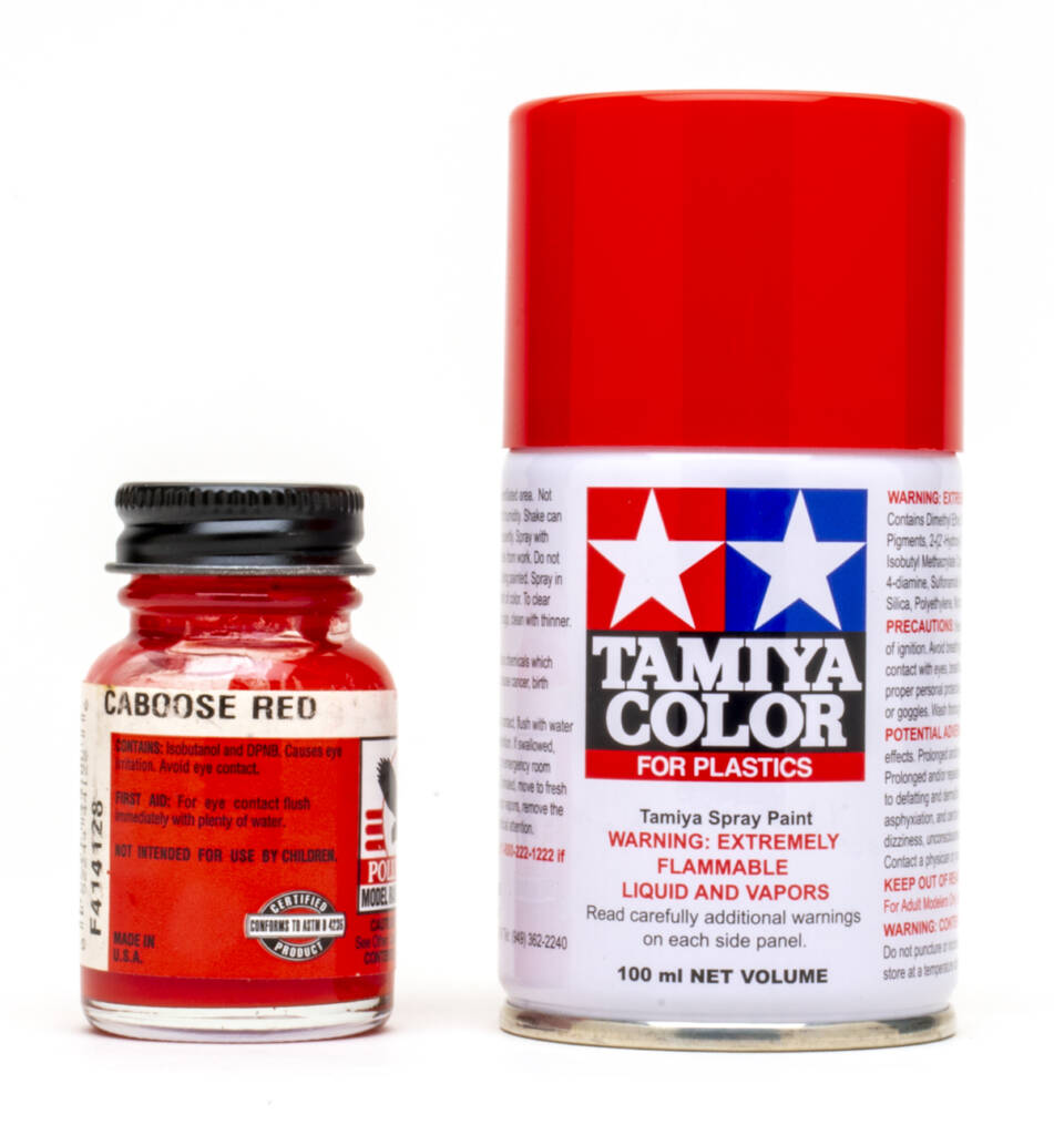 Color photo showing glass bottle of red paint with red, white, and black label and black metal lid and spray can with red, white, blue, and black graphics and red plastic cap on white background.